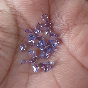 May include: A collection of pear-shaped, light purple gemstones. The gemstones are loose and scattered on a hand.