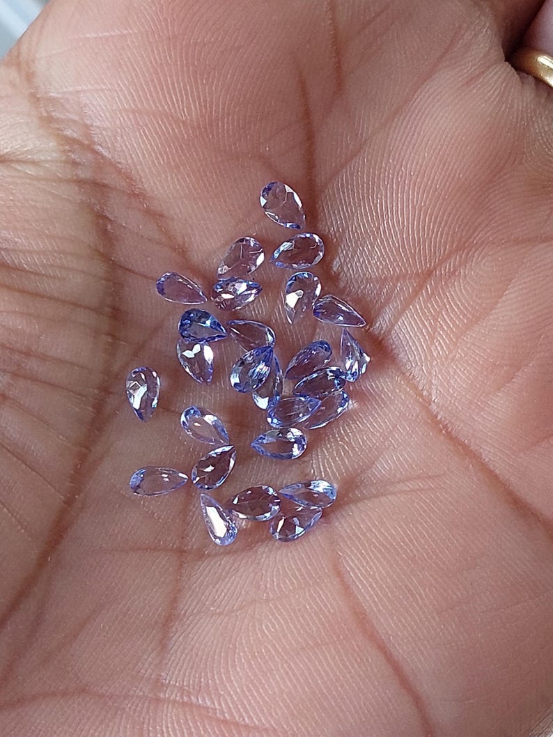 May include: A collection of pear-shaped, light purple gemstones. The gemstones are loose and scattered on a hand.