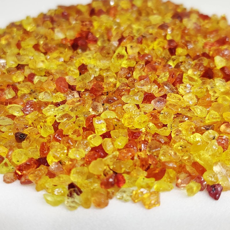 May include: A close-up view of a pile of small, irregularly shaped gemstones. The gemstones are primarily yellow and orange, with some red hues. The stones are scattered, creating a textured surface.