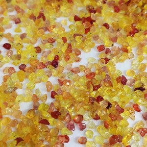 May include: A scattering of small, polished gemstones in shades of yellow, orange, and red. The gems are irregularly shaped and appear to be scattered on a white surface, creating a vibrant and textured display.