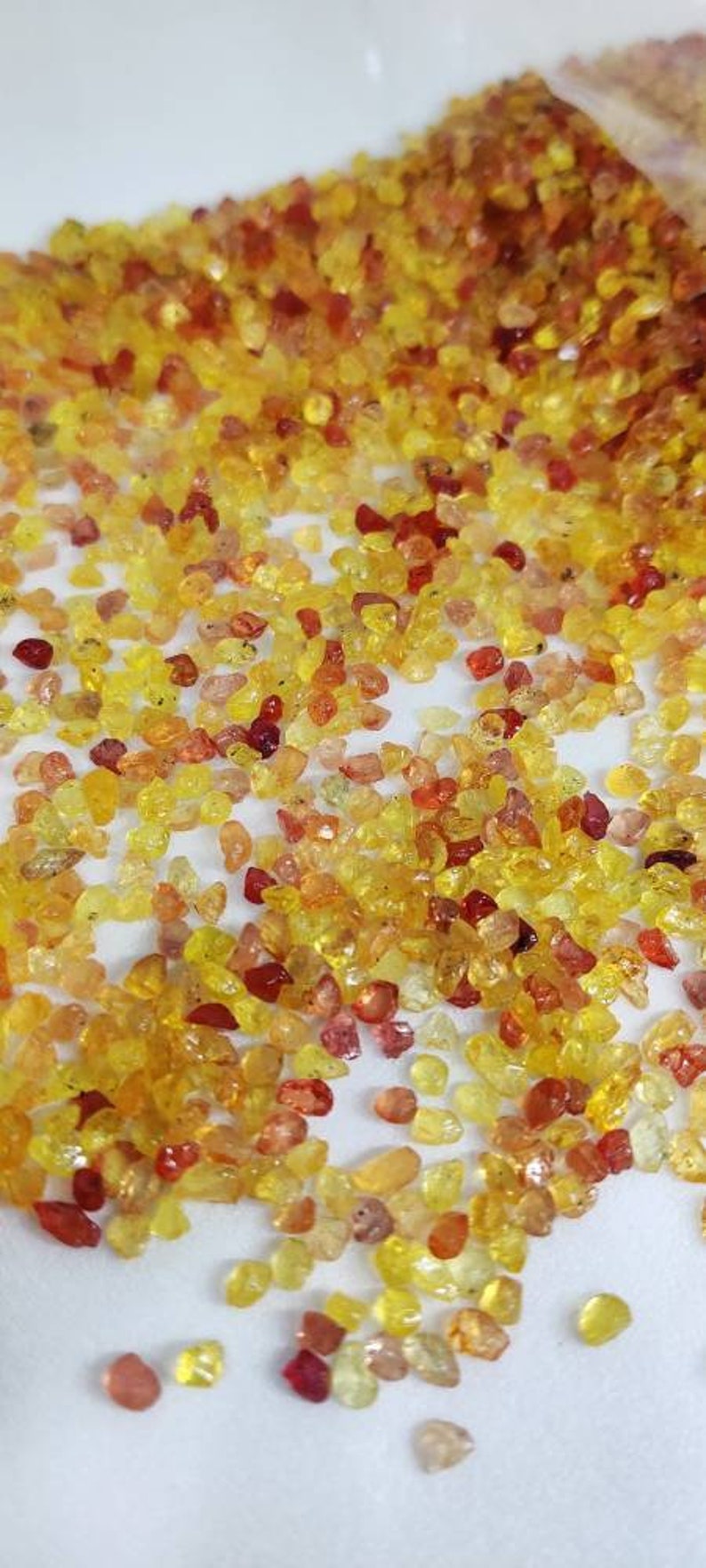 May include: A scattering of small, polished gemstones in shades of yellow, orange, and red. The gems are irregularly shaped and appear to be scattered on a white surface, creating a vibrant and textured display.
