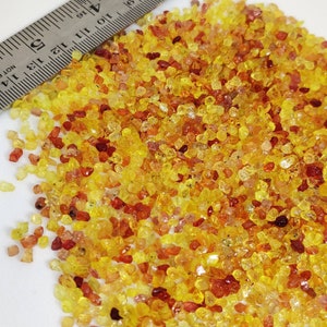 May include: A pile of small, irregularly shaped gemstones in shades of yellow, orange, and red. A metal ruler with inch markings is partially visible, providing scale. The gemstones are scattered across a white surface.