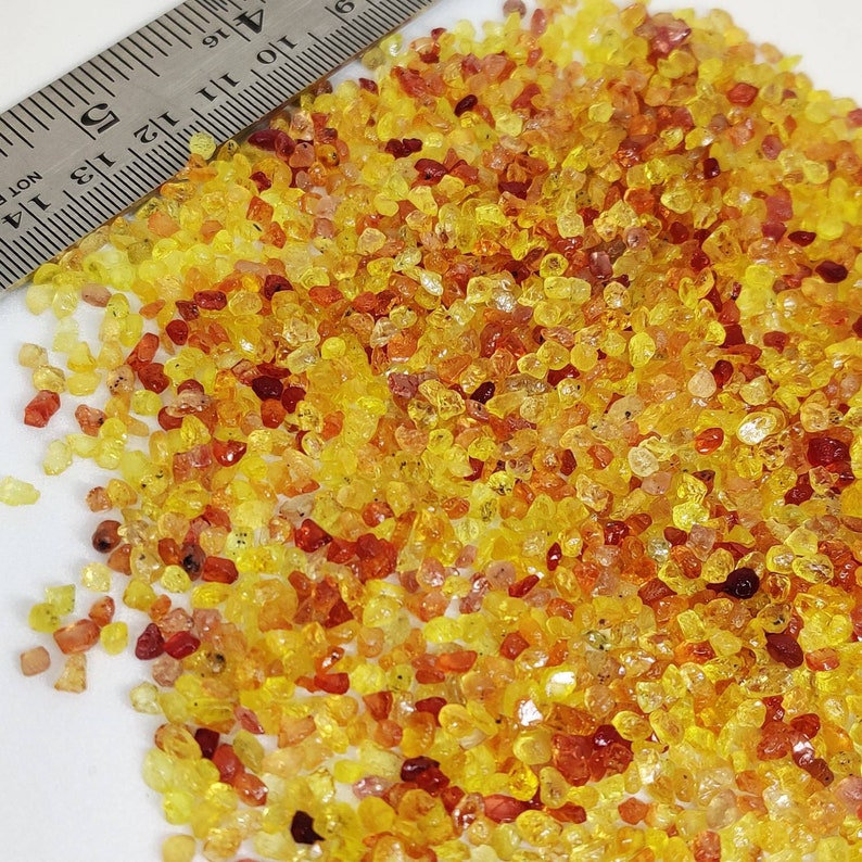 May include: A pile of small, irregularly shaped gemstones in shades of yellow, orange, and red. A metal ruler with inch markings is partially visible, providing scale. The gemstones are scattered across a white surface.