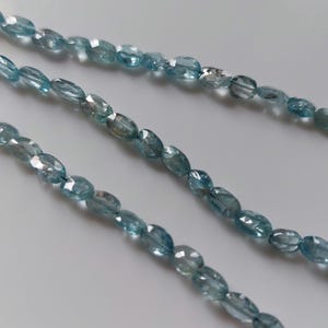 Puede incluir: Un hilo de cuentas de piedras preciosas de color azul claro, con forma ovalada y facetas. Las piedras translúcidas presentan un acabado pulido con múltiples superficies reflectantes, dispuestas en una línea continua que cruza el encuadre.