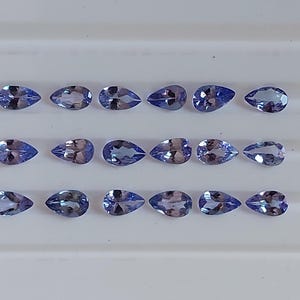 May include: A collection of 18 pear-shaped gemstones, each with a light blue to purple hue. The gemstones are arranged in three rows of six, on a white surface.