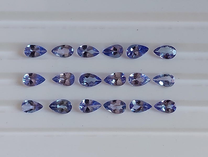 May include: A collection of 18 pear-shaped gemstones, each with a light blue to purple hue. The gemstones are arranged in three rows of six, on a white surface.
