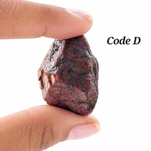 Könnte beinhalten: Ein raues, dunkel rotbraunes Mineralexemplar mit einer texturierten, unebenen Oberfläche und sichtbaren kristallinen Facetten. Der Stein ist mit dem Text Code D beschriftet, der in einer dunklen Serifenschrift vor weißem Hintergrund erscheint.