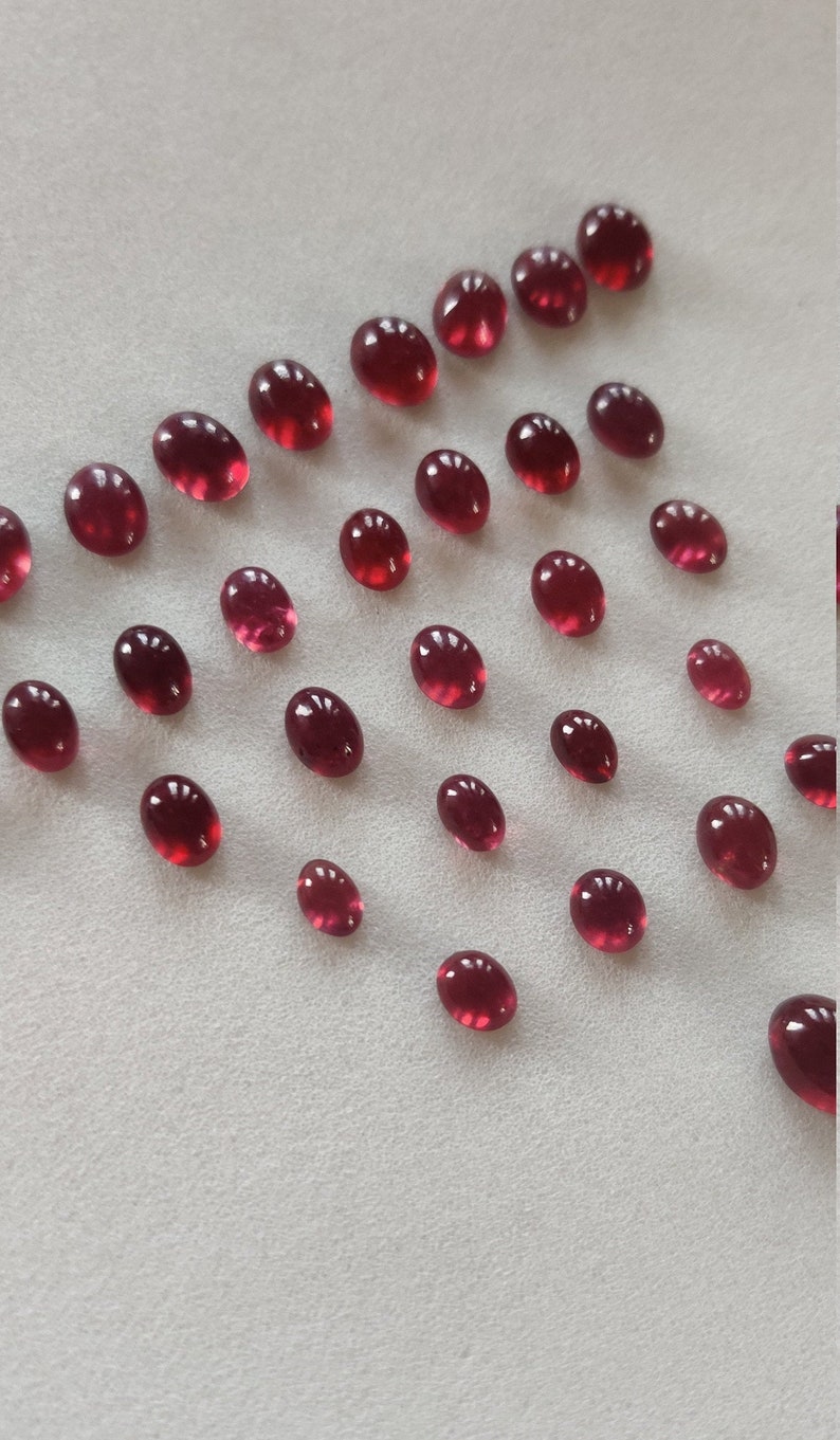 May include: A collection of oval-shaped, red gemstones. The gemstones are scattered on a white surface.