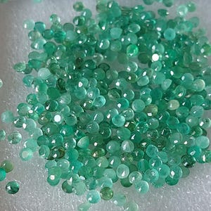 May include: A scattering of small, faceted emerald gemstones in various shades of green. The gemstones are spread across a white surface, with light reflecting off their cut facets. The overall effect is a sparkling, jewel-toned display.