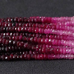 Puede incluir: Una hebra de cuentas de rubí facetadas en un degradado de tonos rojos y rosados. Las cuentas son pequeñas y uniformes en tamaño, creando un aspecto suave y elegante.