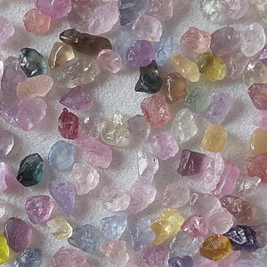 May include: A close-up of a collection of raw gemstones in a variety of colours, including pink, purple, yellow, green, and brown. The irregularly shaped stones are scattered across a white surface, highlighting their natural beauty and varied hues.