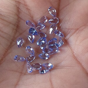 May include: A collection of pear-shaped, faceted, light blue gemstones. The gemstones are scattered on a hand.