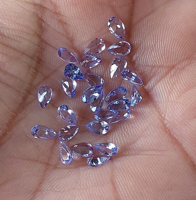 May include: A collection of pear-shaped, faceted, light blue gemstones. The gemstones are scattered on a hand.