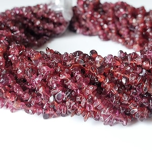 Puede incluir: Un hilo de pequeñas esquirlas de granate de color rojo intenso e irregular. Las cuentas son translúcidas con una superficie pulida y reflectante, dispuestas en un bucle circular para mostrar las formas variadas y los tonos carmesí oscuros.