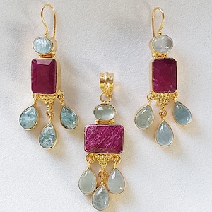 以下が含まれることがあります： 正方形のピンクの宝石と、より小さな涙型の青い宝石がついた、金のイヤリングとペンダントのセットです。