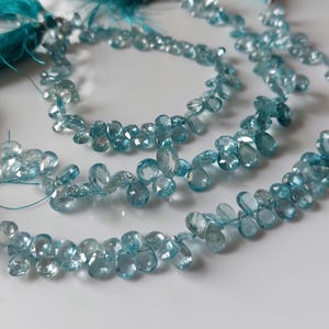 Puede incluir: Múltiples hilos de piedras preciosas de color azul claro, facetadas y con forma de pera, dispuestas en un patrón curvo. Cada hilo está asegurado con una borla de color verde azulado vibrante, con pequeñas superficies claras.