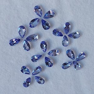 May include: A collection of pear-shaped gemstones in various shades of blue, arranged in a scattered pattern on a white surface.
