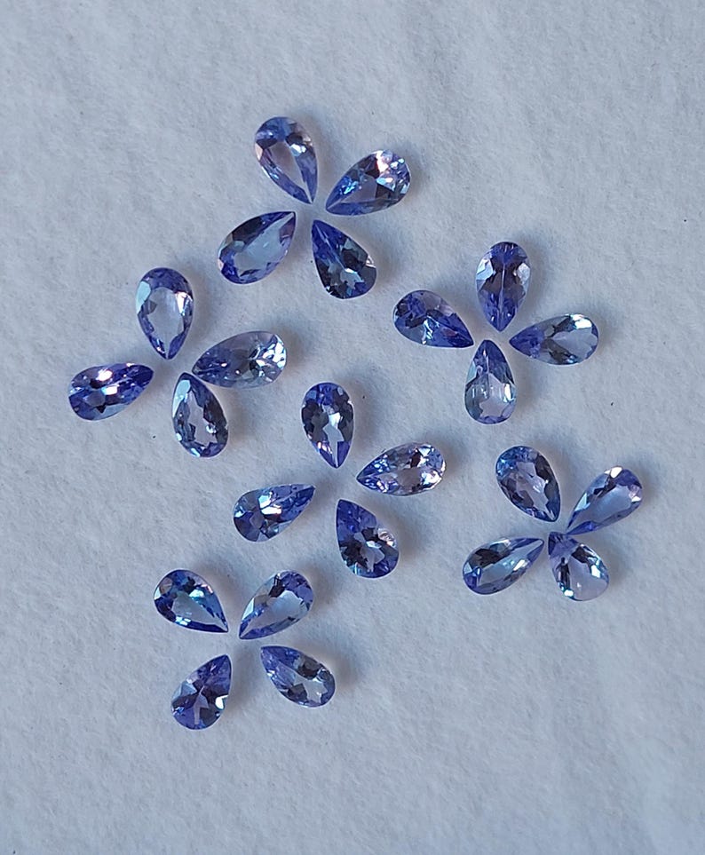May include: A collection of pear-shaped gemstones in various shades of blue, arranged in a scattered pattern on a white surface.