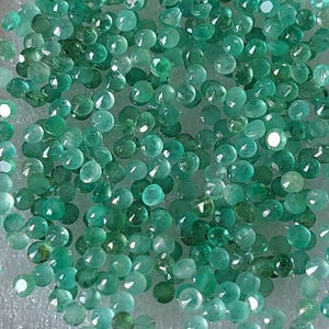 May include: A close-up view of numerous small, round emerald gemstones. The gems are a range of green hues, from light to dark, and have multiple facets. The light catches the cut surfaces, creating a sparkling effect. The image showcases the natural beauty of the emeralds.
