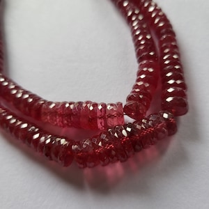 May include: A strand of red gemstone beads, faceted and strung on a thread. The beads are a deep red colour and have a slightly irregular shape.