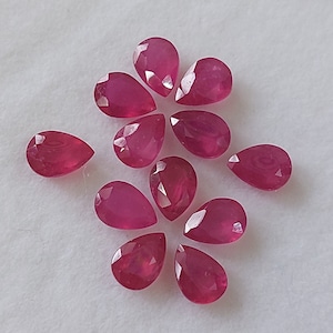 May include: Ten pear-shaped ruby gemstones, each with a deep red hue and a faceted cut. The gemstones are arranged in a loose, organic pattern on a white surface.
