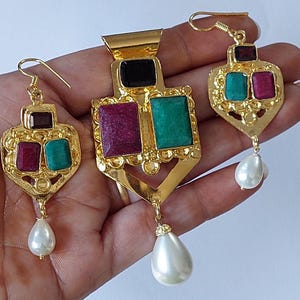 May include: A gold-coloured jewellery set comprising a pendant and matching earrings. The pendant and earrings feature ruby, emerald, and black-coloured stones. Each piece has a teardrop-shaped pearl pendant.