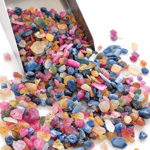 Può includere: Un paletta in metallo versa una collezione di piccole pietre preziose grezze e multicolori su una superficie bianca. Le pietre sono traslucide e opache, con varie tonalità di blu, rosa, giallo e colori chiari.
