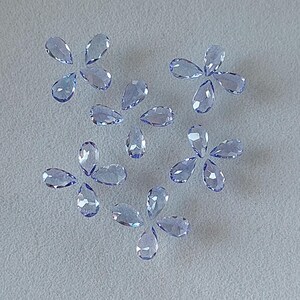 May include: A collection of twelve pear-shaped faceted gemstones in a light blue color. The gemstones are arranged in a scattered pattern on a white surface.