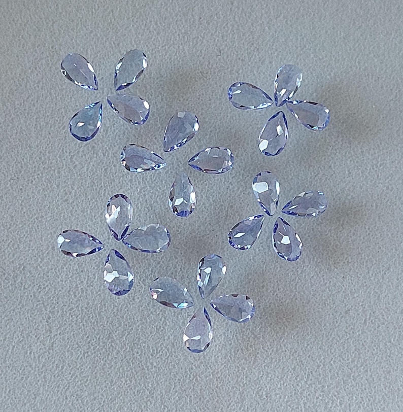 May include: A collection of twelve pear-shaped faceted gemstones in a light blue color. The gemstones are arranged in a scattered pattern on a white surface.