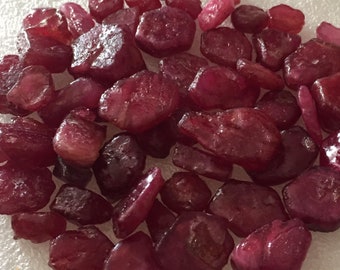 May include: A collection of raw, uncut ruby gemstones. The stones vary in size and shape, displaying a deep red hue with some areas appearing darker. The gems are scattered on a white surface, showcasing their natural, unpolished state.