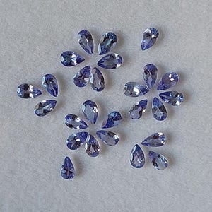 May include: A collection of pear-shaped gemstones in various shades of blue, arranged in a decorative pattern on a white surface.