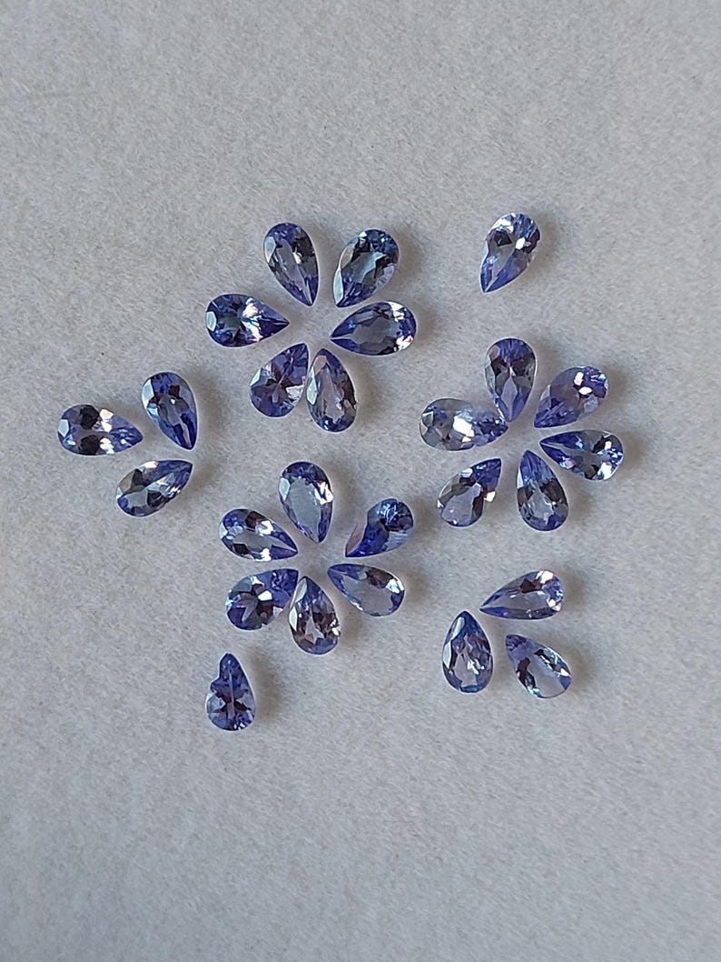 May include: A collection of pear-shaped gemstones in various shades of blue, arranged in a decorative pattern on a white surface.