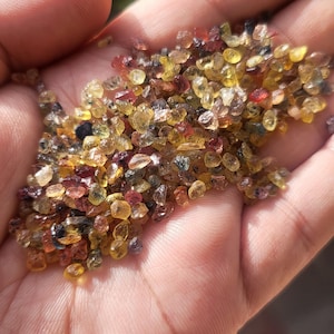 May include: A close-up of a collection of small, uncut gemstones. The gems are a mix of colours, including yellow, orange, red, and blue. The stones are irregularly shaped and held in a hand. The image is well-lit, showing the gems' details.