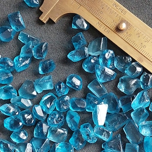 May include: A close-up of a collection of small, faceted, blue gemstones. The gemstones are scattered on a dark gray surface. A brass ruler is visible in the background.