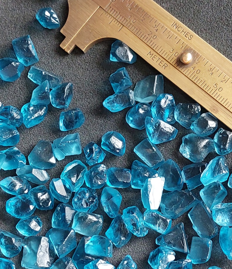 May include: A close-up of a collection of small, faceted, blue gemstones. The gemstones are scattered on a dark gray surface. A brass ruler is visible in the background.