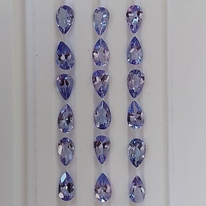 May include: A collection of 18 pear-shaped gemstones, arranged in three rows of six. The gemstones are a light purple color with a slight blue hue.