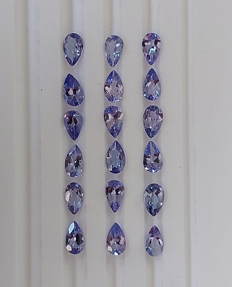 May include: A collection of 18 pear-shaped gemstones, arranged in three rows of six. The gemstones are a light purple color with a slight blue hue.