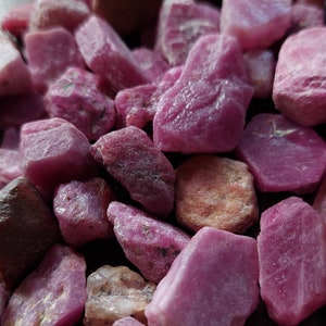 May include: A close-up of a pile of pink ruby gemstones. The rough, unpolished stones are various sizes and shapes.