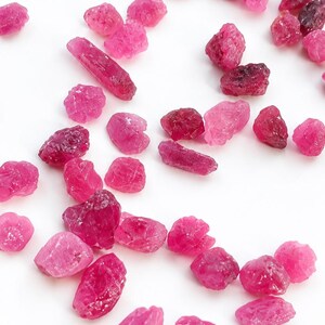 Puede incluir: Una colección de pequeñas piedras preciosas de rubí en bruto y sin pulir esparcidas sobre una superficie blanca lisa. Las piedras muestran varios tonos de rosa vibrante y rojo intenso, con formas irregulares y texturizadas.