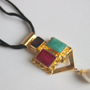 May include: Gold-toned pendant necklace with a black cord. The pendant features a geometric design with three colorful gemstones: red, teal, and a dark red. A white teardrop-shaped pearl hangs from the bottom.