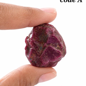 Puede incluir: Una piedra preciosa de rubí en bruto de color magenta intenso con una textura superficial rugosa, irregular y facetada. La piedra muestra inclusiones naturales y depósitos minerales oscuros en toda su estructura. El texto code A es visible arriba a la derecha.