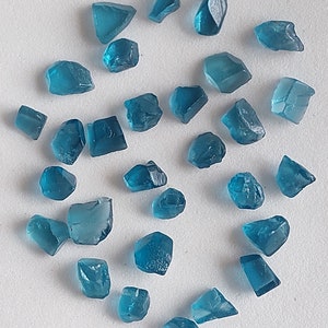 May include: A collection of teal blue gemstone chips, some with a more rectangular shape and others with a more irregular shape. The chips are scattered on a white surface.