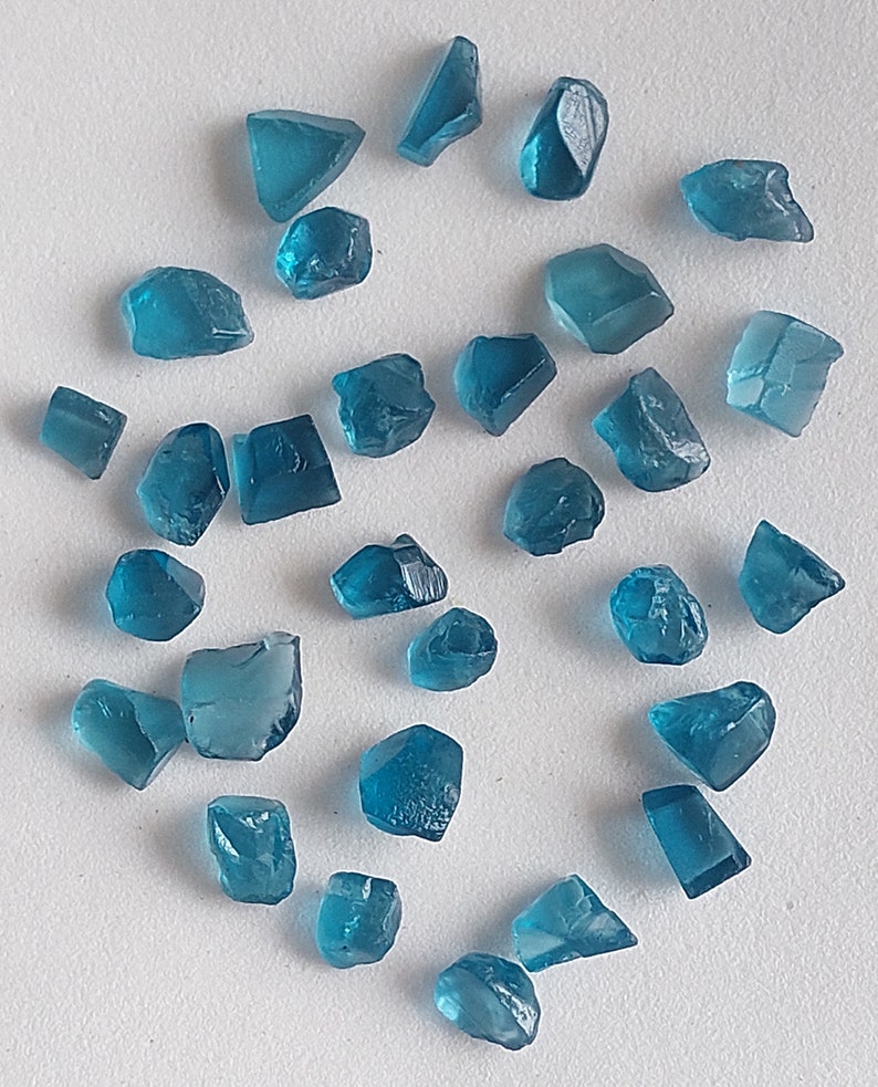 May include: A collection of teal blue gemstone chips, some with a more rectangular shape and others with a more irregular shape. The chips are scattered on a white surface.