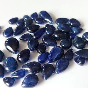 Puede incluir: Una colección de piedras preciosas de cianita azul sueltas, pulidas y con forma de lágrima. Las piedras muestran tonos variados de azul marino profundo y azul real con superficies lisas y reflectantes e inclusiones naturales visibles.