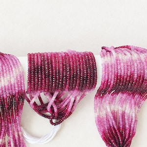 May include: Three strands of faceted gemstone beads in varying shades of pink, magenta, and deep burgundy. The beads are arranged in a gradient pattern, transitioning from light translucent pink to dark saturated red, strung on thin white cord.