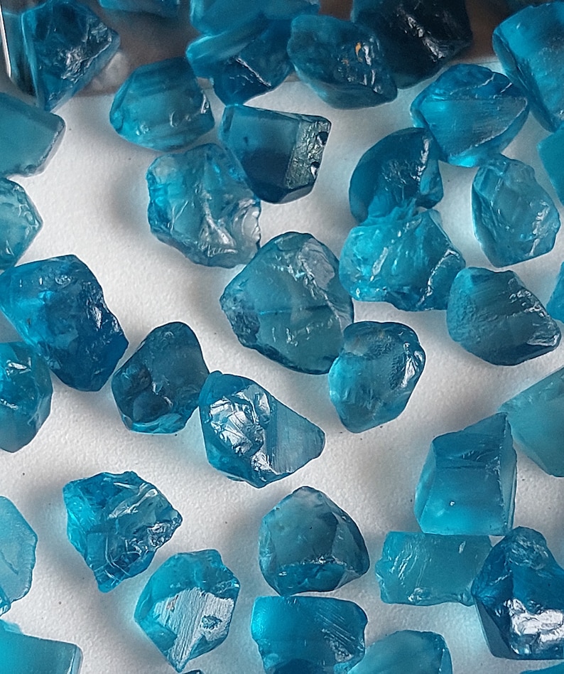 May include: A collection of teal blue gemstone roughs. The rough gemstones are various sizes and shapes. The gemstones are on a white surface.