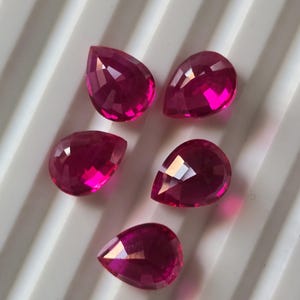 May include: Five faceted, pear-shaped gemstones in a vibrant magenta hue. The gems are arranged on a white, ridged surface, showcasing their brilliant cut and reflective qualities. These gemstones are ideal for jewelry making.