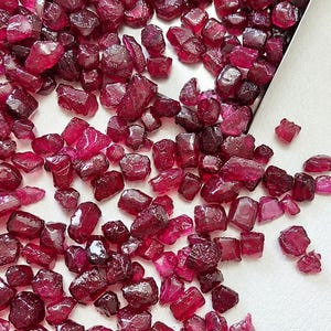 May include: A collection of small, raw, deep red garnet gemstones scattered across a surface, with a portion of the stones contained within a rectangular metal scoop. The stones have irregular, faceted shapes and a glossy, translucent appearance.