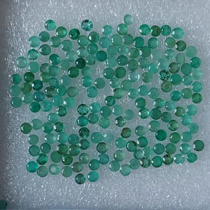 May include: Numerous small, round, faceted emerald gemstones in shades of green and teal. The gemstones are displayed on a white, textured surface, highlighting their polished facets and natural colour variations.