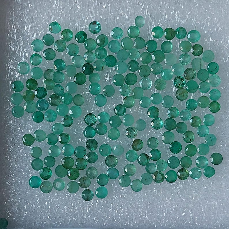 May include: Numerous small, round, faceted emerald gemstones in shades of green and teal. The gemstones are displayed on a white, textured surface, highlighting their polished facets and natural colour variations.